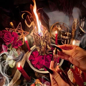 May include: A mystical scene featuring a lit gold candle with a detailed figure, surrounded by red roses, candles, and a small vial held by a person. The image has a dark, atmospheric aesthetic with a focus on ritualistic elements.