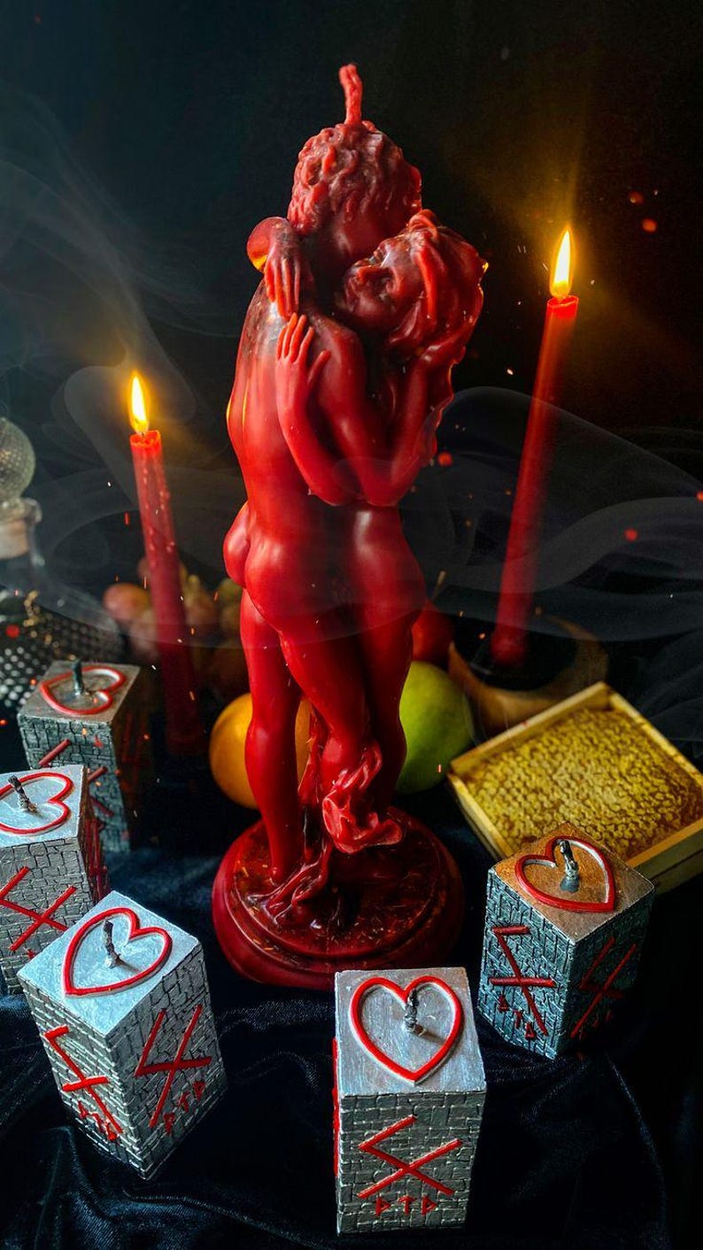 K&ouml;nnte beinhalten: Eine rote Kerze in Form von zwei sich umarmenden Figuren ist der Mittelpunkt, umgeben von brennenden roten Kerzen und dekorativen W&uuml;rfeln mit Herz- und Kreuzmotiven. Die Szene spielt sich vor einem dunklen Hintergrund ab.