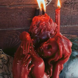 K&ouml;nnte beinhalten: Eine rote Kerzenskulptur, die verschlungene Figuren darstellt, mit zwei brennenden Dochten. Die Kerze steht vor einem Holzgrund, mit einem dekorativen metallenen Halbmond dar&uuml;ber. Das Wachs hat ein strukturiertes, geschmolzenes Aussehen.