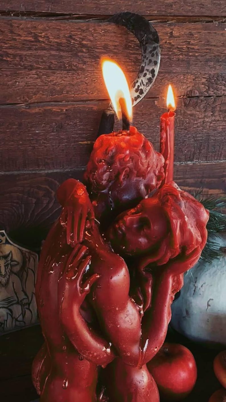 K&ouml;nnte beinhalten: Eine rote Kerzenskulptur, die verschlungene Figuren darstellt, mit zwei brennenden Dochten. Die Kerze steht vor einem Holzgrund, mit einem dekorativen metallenen Halbmond dar&uuml;ber. Das Wachs hat ein strukturiertes, geschmolzenes Aussehen.