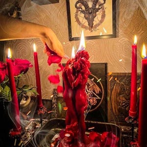 K&ouml;nnte beinhalten: Eine rote Kerzenskulptur mit zwei sich umarmenden Figuren, umgeben von roten Kerzen und Rosenbl&auml;ttern. Eine Hand h&auml;lt eine Rose, was die rituelle Szene verst&auml;rkt. Im Hintergrund ist ein gerahmtes Bild eines Ziegenkopfes zu sehen, zusammen mit anderen okkulten Symbolen.