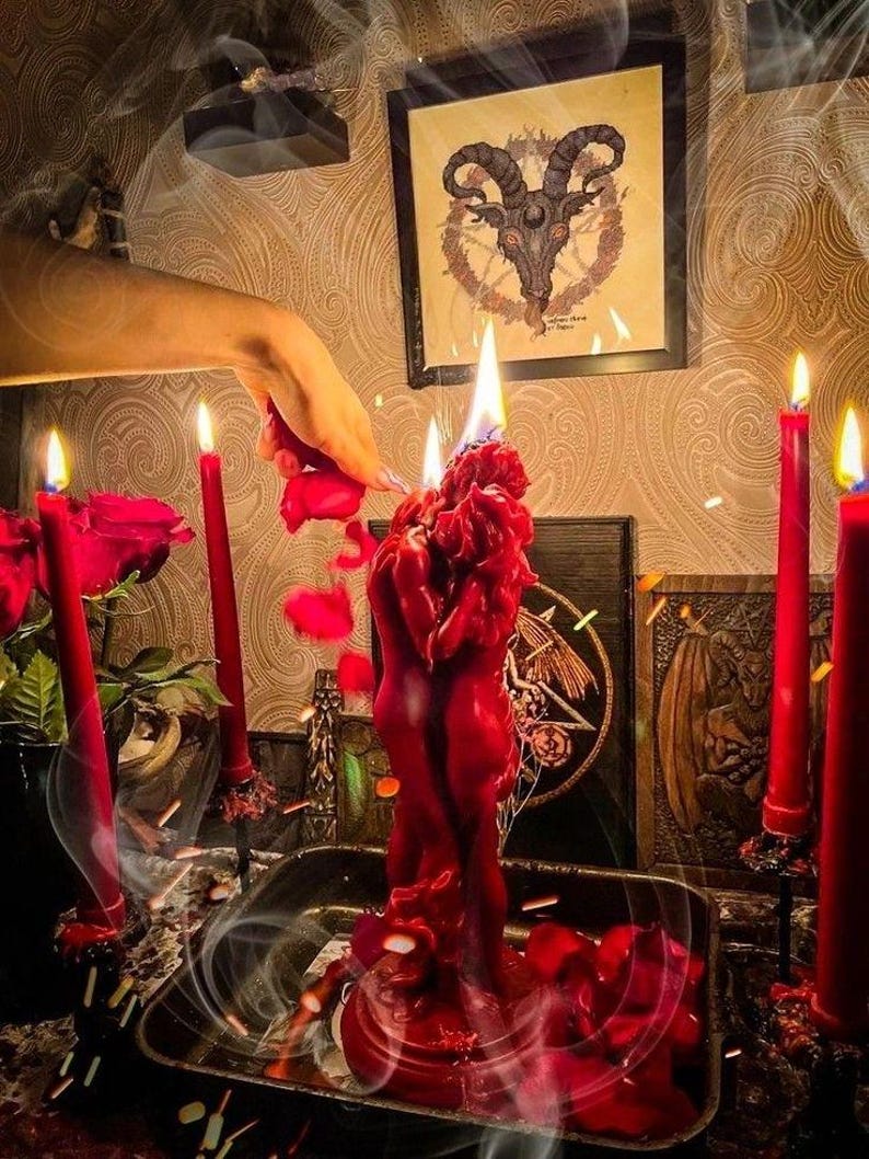 K&ouml;nnte beinhalten: Eine rote Kerzenskulptur mit zwei sich umarmenden Figuren, umgeben von roten Kerzen und Rosenbl&auml;ttern. Eine Hand h&auml;lt eine Rose, was die rituelle Szene verst&auml;rkt. Im Hintergrund ist ein gerahmtes Bild eines Ziegenkopfes zu sehen, zusammen mit anderen okkulten Symbolen.