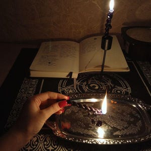 May include: A lit black spiral candle on a silver tray with a burning wick. An open book and a second lit candle are in the background. The scene is dimly lit, creating a mystical atmosphere.
