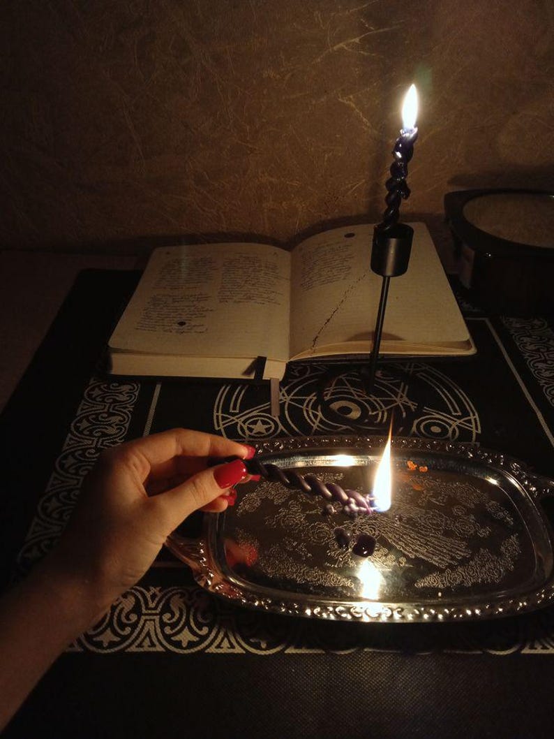 May include: A lit black spiral candle on a silver tray with a burning wick. An open book and a second lit candle are in the background. The scene is dimly lit, creating a mystical atmosphere.