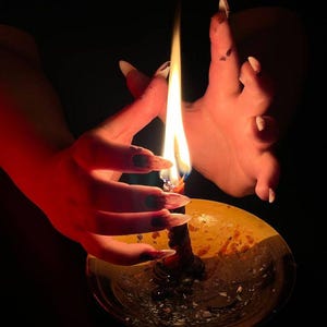 May include: A lit candle in a gold-colored holder. The flame is bright against the dark background. The candle is surrounded by a hand with long, pointed nails.