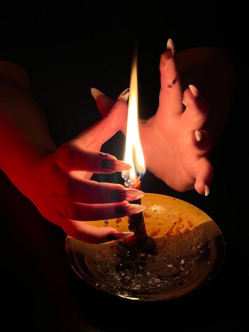 May include: A lit candle in a gold-colored holder. The flame is bright against the dark background. The candle is surrounded by a hand with long, pointed nails.