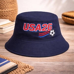 May include: A navy blue bucket hat featuring the text "USA26" in red and white, and a soccer ball graphic. The hat has a wide brim and is made of a durable fabric. Ideal for sports fans.