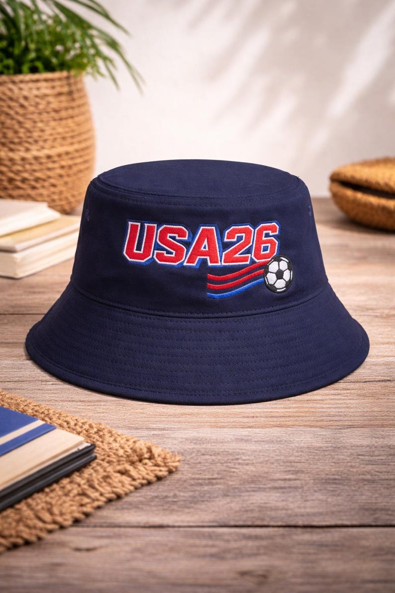 May include: A navy blue bucket hat featuring the text "USA26" in red and white, and a soccer ball graphic. The hat has a wide brim and is made of a durable fabric. Ideal for sports fans.