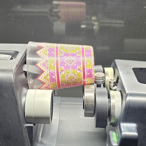 May include: A black eufy Make rotary attachment machine holding a clear glass tumbler. The tumbler features a vibrant pink, yellow, and white geometric pattern with intricate circular and triangular designs wrapped around its exterior surface.