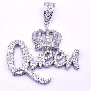 May include: A silver pendant with the word "Queen" spelled out in cursive, encrusted with small, sparkling stones. Above the word is a crown, also covered in the same stones. The pendant has a loop at the top for a chain.