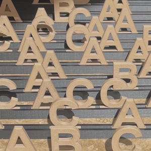 Pode incluir: Uma coleção de letras A, B e C de madeira em tom castanho claro, dispostas em várias filas. Cada letra apresenta uma tipografia sem serifa limpa e um acabamento mate suave, concebida para ficar de pé numa superfície plana.