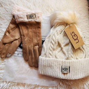 May include: A set of winter accessories, including tan gloves and a cream-colored cable-knit beanie with a faux fur pom-pom. Both items feature the "UGG Australia" logo. The gloves are a light brown suede-like material.
