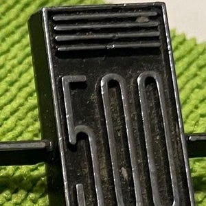 May include: A rectangular black metal emblem featuring the number 500 in a stylised, raised silver font. Above the number, there are three horizontal silver bars. The emblem has a metallic finish and a clean, geometric design with sharp edges.
