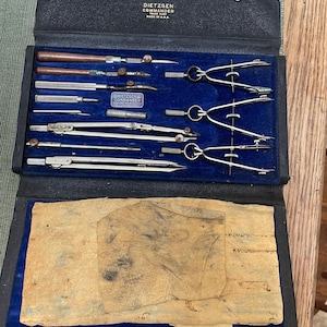 May include: Open black case with a blue velvet interior containing a set of vintage drafting tools. The tools include compasses, dividers, and other instruments. The case is labeled "DIETZGEN COMMANDER" and "MADE IN U.S.A."