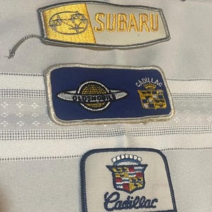 May include: A collection of embroidered patches featuring car brand logos. Includes Lexus, Subaru, Oldsmobile, and Cadillac patches. The Cadillac patches include the word "SERVICE". The patches are in various colors, including black, gold, blue, and white.