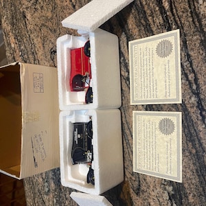 May include: Two miniature vintage car models, one red and one black, are displayed in protective white foam packaging. Accompanying the cars are two certificates of authenticity from the National Motor Museum Mint.