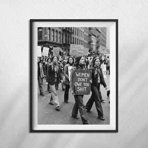 May include: Framed black and white print depicting a protest march. A group of people are walking down a city street, some holding signs. One sign reads "WOMEN DON'T OWE YOU SHIT". The image is a historical photograph.