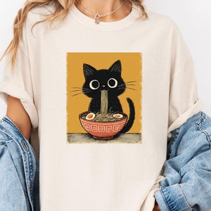 May include: Cream-colored t-shirt featuring a black cat enjoying ramen from a red bowl. The cat has large eyes and is surrounded by noodles and eggs. The graphic is set against a mustard-yellow background.