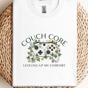 May include: White t-shirt with a graphic of a video game controller decorated with flowers and vines. The text "COUCH CORE" is above the controller, and "LEVELING UP MY COMFORT" is below it.