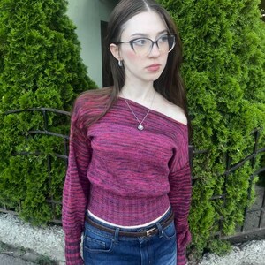 May include: A long-sleeved, off-the-shoulder sweater in a variegated magenta and black knit pattern, paired with a dark blue denim mini skirt featuring a brown leather belt with a gold-toned buckle and a silver pendant necklace.