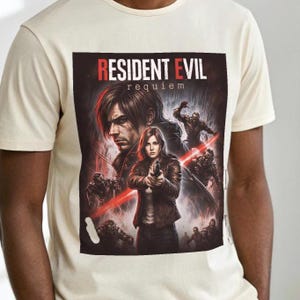 May include: Cream-colored t-shirt featuring a graphic from the video game "Resident Evil: Requiem." The design includes characters with guns and zombies, with the game's title in red. The shirt is made of a soft material.
