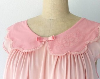 May include: A pink, short-sleeved nightgown with a scalloped lace collar and a bow at the neckline. The gown is made of a soft, sheer fabric.
