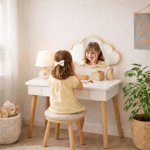 May include: A white vanity table with wooden legs and a cloud-shaped mirror. A young person in a yellow dress sits at the vanity, looking in the mirror. A lamp, a small stool, and a woven basket are also visible.