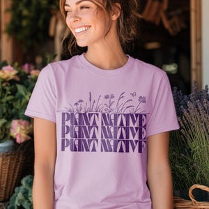 May include: A light purple t-shirt with the word "PLANT NATIVE" in a stacked design. The text is in a darker purple and is above a line of stylized plants and a butterfly.