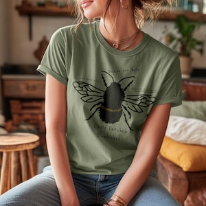 May include: Olive green t-shirt featuring a black illustration of a bumblebee with the text "Needs our help" and "Rusty Patched Bumblebee." The shirt also includes the phrases "Cool Personality" and "Federally Endangered."