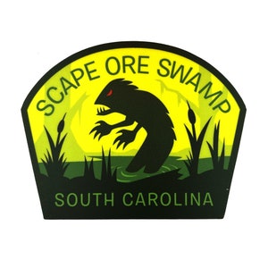 May include: A patch with a black silhouette of a swamp creature with red eyes, set against a yellow background. The text "SCAPE ORE SWAMP" arches across the top, and "SOUTH CAROLINA" is at the bottom. The design includes cattails and water.