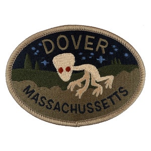 May include: Embroidered patch with a green, brown, and black background. The patch features a white alien with red eyes walking across a field. The text "DOVER MASSACHUSETTS" is embroidered in brown on the patch.