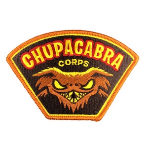 May include: Embroidered patch featuring a stylized Chupacabra design. The patch has a black background with the words "CHUPACABRA CORPS" in yellow and red. The Chupacabra is depicted with yellow eyes and a menacing expression, outlined in brown.