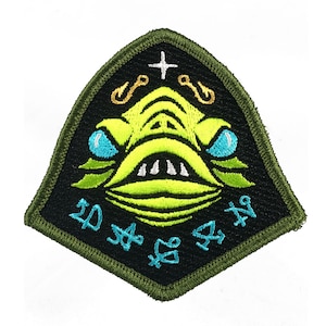 Puede incluir: Parche bordado con un rostro de criatura verde lima estilizado con ojos azules, dientes blancos y una boca ancha. El parche tiene un fondo negro con detalles turquesa y dorados, y un borde verde. El parche tiene texto.