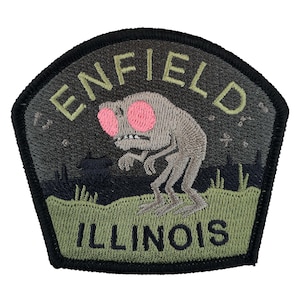 Puede incluir: Un parche bordado en verde, negro y gris con el texto "ENFIELD ILLINOIS" y una criatura de dibujos animados con ojos rosas.