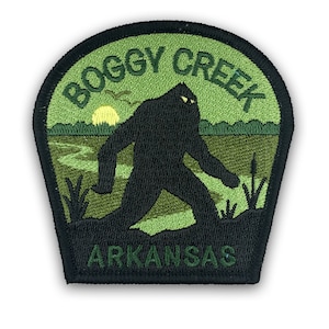 May include: Green and black embroidered patch featuring a silhouette of Bigfoot walking through a swamp. The text "BOGGY CREEK ARKANSAS" is embroidered around the image.