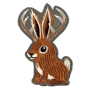 May include: Embroidered patch featuring a brown rabbit with large ears and a gray background. The rabbit has detailed fur and a friendly expression. The patch is designed for application on clothing or accessories.