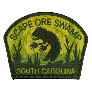 May include: Embroidered patch featuring a dark green swamp creature with red eyes, set against a green and yellow background. The patch reads "SCAPE ORE SWAMP" and "SOUTH CAROLINA" in green lettering. The patch has a dark green border.