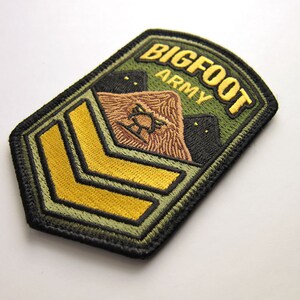 May include: Embroidered patch with the words "BIGFOOT ARMY" in gold lettering on a green background. The patch features a brown Bigfoot illustration and two black silhouettes. Gold chevrons are at the bottom.
