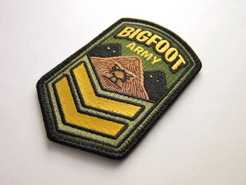 May include: Embroidered patch with the words "BIGFOOT ARMY" in gold lettering on a green background. The patch features a brown Bigfoot illustration and two black silhouettes. Gold chevrons are at the bottom.