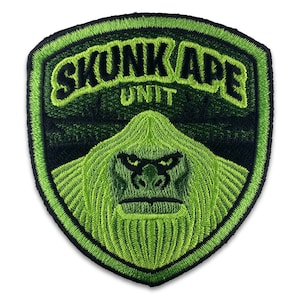 Könnte beinhalten: Grün-schwarzer gestickter Aufnäher mit dem Text "Skunk Ape Unit" in geschwungener Schrift. In der Mitte des Aufnähers befindet sich ein grünes, stilisiertes Bild eines Gorillagesichts.