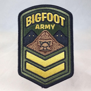 May include: Embroidered patch with the words "BIGFOOT ARMY" in gold lettering. The patch features a brown Bigfoot face, two dark blue mountains, and two yellow chevrons on a green background, with a black border.