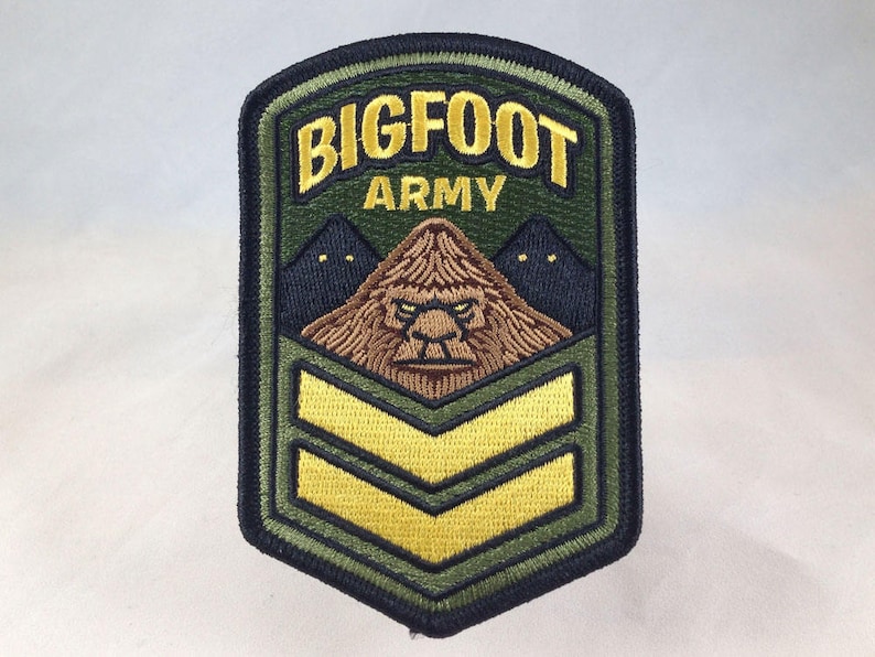 May include: Embroidered patch with the words "BIGFOOT ARMY" in gold lettering. The patch features a brown Bigfoot face, two dark blue mountains, and two yellow chevrons on a green background, with a black border.