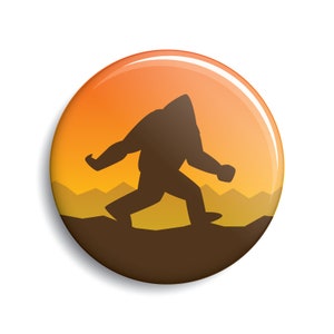 Puede incluir: Un botón redondo brillante con una silueta de Bigfoot caminando sobre un paisaje marrón. El fondo transiciona de amarillo a naranja, con siluetas de montañas.