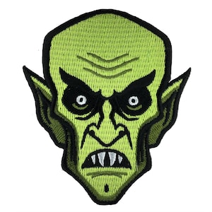 Puede incluir: Parche bordado verde de la cara de un monstruo con orejas grandes, ojos negros y dientes afilados.