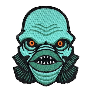 May include: Green and black embroidered patch of a creature from the black lagoon with orange eyes.