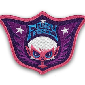 Puede incluir: Parche bordado morado y rosa con una cara de hada de dibujos animados y el texto "Fairy Force" en una fuente azul y blanca.