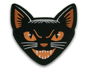 Op de afbeelding: Zwart kattengezicht met oranje ogen en tanden, geborduurd op een zwarte stoffen patch.