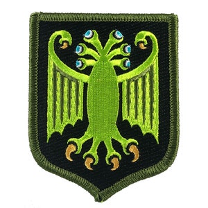 May include: Embroidered patch featuring a green, stylized creature with multiple eyes and clawed feet, set against a black shield shape. The patch has a green border and is likely for clothing or accessories.