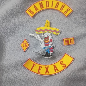 Puede incluir: Parches bordados con las palabras "BANDIDOS" y "TEXAS" en pancartas arqueadas amarillas. Un personaje de dibujos animados con sombrero sostiene una pistola y un cuchillo. También incluye parches "1%" y "MC".