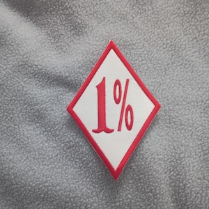 Puede incluir: Un parche blanco en forma de rombo con borde rojo y el texto "1%" en rojo. El parche está sobre una superficie gris texturizada.